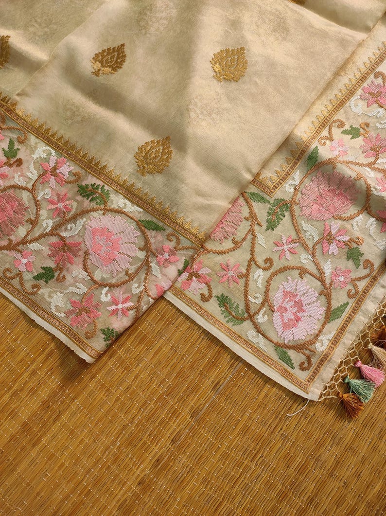 Pode incluir: T&ecirc;xtil creme com padr&otilde;es de folhas douradas e bordados florais em rosa, verde e castanho. A borda apresenta intrincados desenhos florais e uma borda dourada. Tassels em v&aacute;rias cores s&atilde;o vis&iacute;veis.