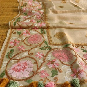 Pode incluir: T&ecirc;xtil bege com bordados florais intrincados em rosa, branco e verde. A borda apresenta um design detalhado, e a borda &eacute; adornada com borlas coloridas. O tecido est&aacute; dobrado, mostrando sua textura e padr&atilde;o.