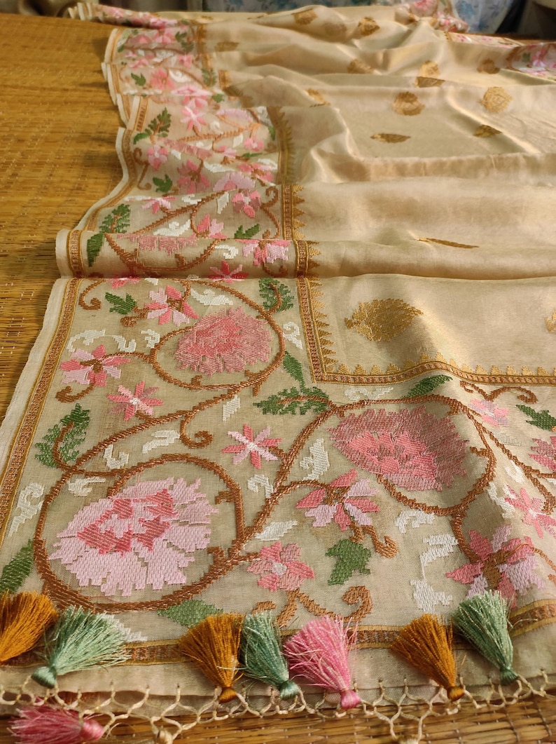 Pode incluir: T&ecirc;xtil bege com bordados florais intrincados em rosa, branco e verde. A borda apresenta um design detalhado, e a borda &eacute; adornada com borlas coloridas. O tecido est&aacute; dobrado, mostrando sua textura e padr&atilde;o.