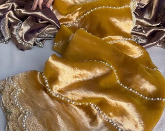 May include: A golden yellow saree with a pearl trim and a delicate lace pattern. The saree is draped over a person's arm and leg, showcasing the fabric's texture and sheen.