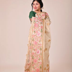 Pode incluir: Um sari creme com bordados florais intrincados em rosa e dourado. O sari apresenta uma borda detalhada com borlas e &eacute; drapeado sobre uma pessoa vestindo um top verde. O design geral &eacute; elegante e tradicional.