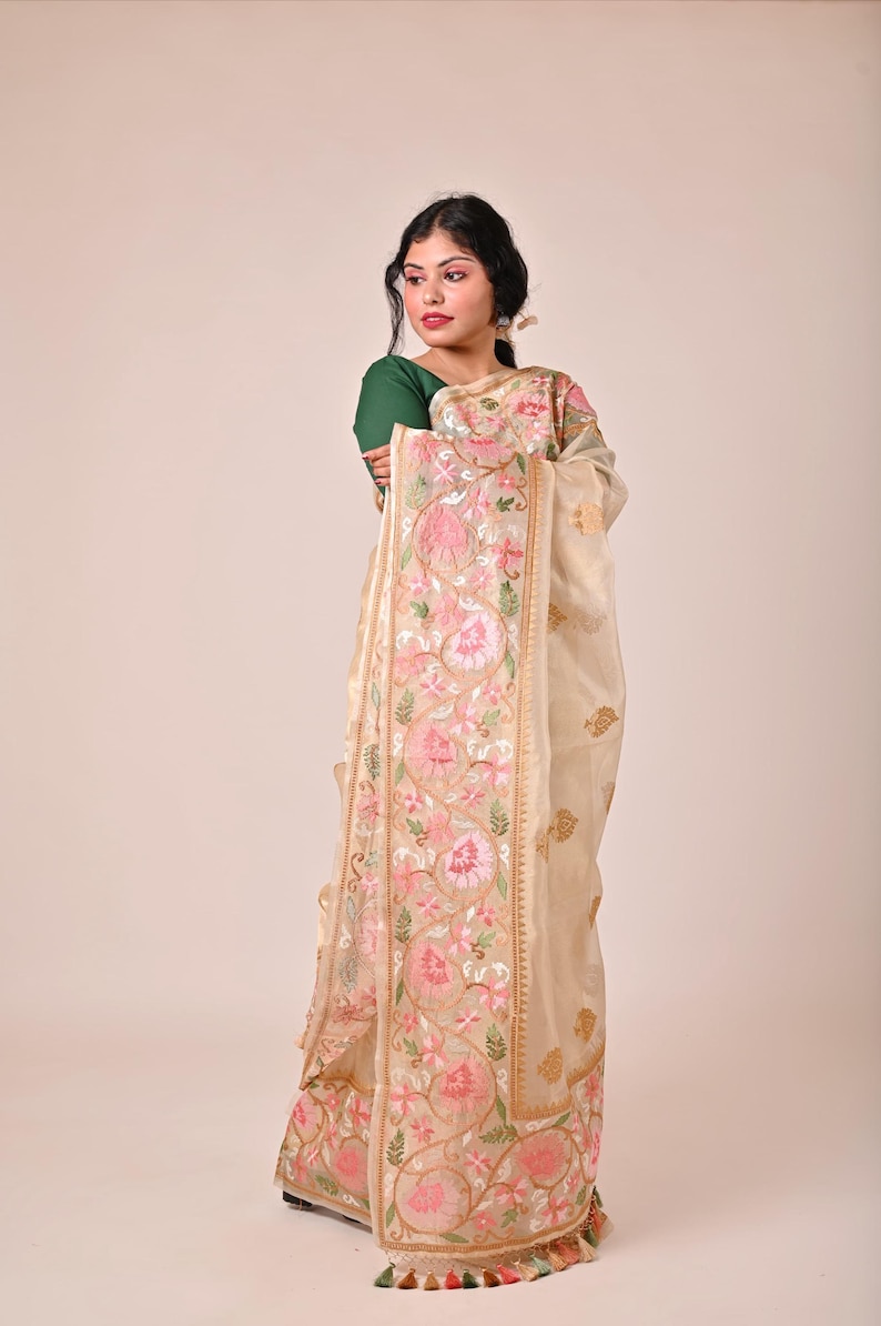 Pode incluir: Um sari creme com bordados florais intrincados em rosa e dourado. O sari apresenta uma borda detalhada com borlas e &eacute; drapeado sobre uma pessoa vestindo um top verde. O design geral &eacute; elegante e tradicional.