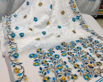 Può includere: Sari bianco con ricamo floreale blu e giallo. Il sari ha una base bianca con un motivo floreale blu e giallo. Il sari è decorato con un bordo di fiori blu e gialli.
