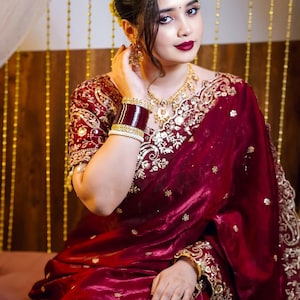 May include: A deep maroon saree featuring intricate gold embroidery along the borders and scattered floral motifs. The garment is paired with a matching short-sleeved blouse, gold jewelry including a necklace and earrings, and wide bangles.