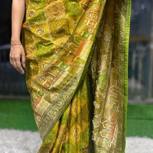 May include: A vibrant olive green saree featuring intricate gold zari borders and a detailed bandhani tie-dye pattern. The fabric displays a complex geometric and floral motif design with a wide, ornate metallic gold hem and a matching pallu.