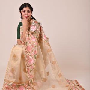 Pode incluir: Um sari bege com bordados florais intrincados em rosa e verde. O sari apresenta uma borda detalhada e detalhes dourados, com um design floral correspondente no pallu. O tecido parece transparente e leve, com uma borda de borlas.