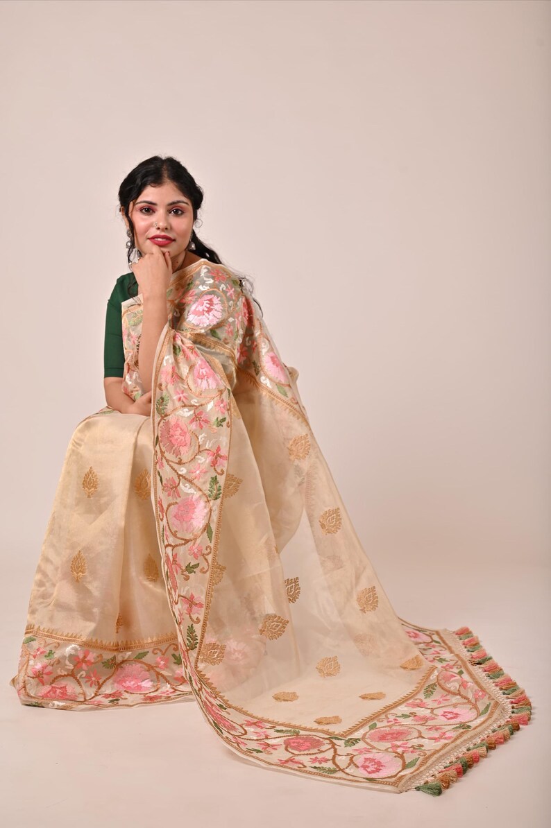 Pode incluir: Um sari bege com bordados florais intrincados em rosa e verde. O sari apresenta uma borda detalhada e detalhes dourados, com um design floral correspondente no pallu. O tecido parece transparente e leve, com uma borda de borlas.