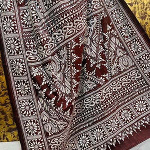 May include: A maroon textile with a white floral and geometric pattern. The fabric is draped, showcasing the intricate design. The pattern includes stylized flowers, leaves, and geometric shapes, creating a detailed and ornate appearance. The textile has a border with a repeating floral design.