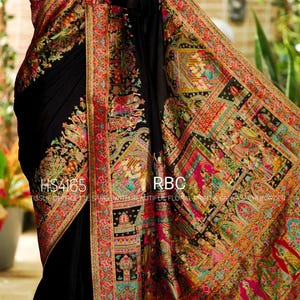 May include: A black saree featuring a vibrant, multi-colored border with intricate floral and traditional patterns. The fabric displays the text HS4165 RBC TISSUE GEORGETTE SAREE WITH BEAUTIFUL FLORAL PRINT & SABYASACHI BORDER. A gold necklace is visible at the neck.