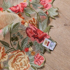 May include: Close-up of a sheer fabric with intricate floral embroidery. The design features large roses in shades of pink, orange, and red, with green leaves and small yellow flowers. A tag is visible on the edge.