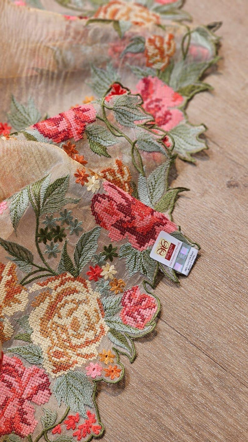 May include: Close-up of a sheer fabric with intricate floral embroidery. The design features large roses in shades of pink, orange, and red, with green leaves and small yellow flowers. A tag is visible on the edge.