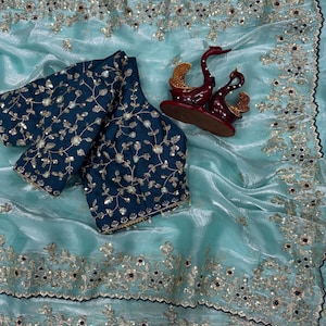 May include: A light blue saree with a dark blue blouse, both featuring intricate gold embroidery and small mirror accents. The saree has a wide decorative border with floral patterns, and a dark red swan-shaped decorative ornament is placed on top.