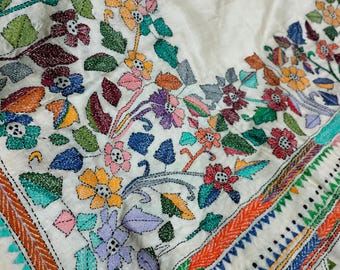 May include: A white cotton saree with a colorful floral embroidery design. The embroidery features a variety of flowers, leaves, and vines in shades of pink, blue, green, yellow, and red. The saree also has a border with a geometric pattern in shades of orange, red, green, and blue.