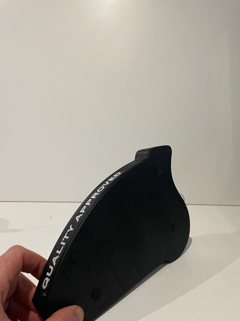 May include: A black, curved object with the text "QUALITY APPROVED" in white lettering. The object has a rounded shape with a small, raised section on one end and three small, circular feet on the bottom.