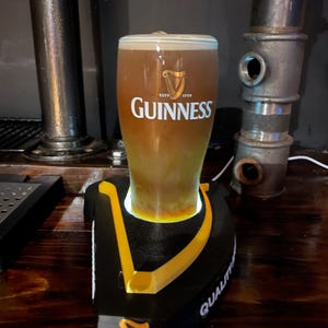May include: A pint of Guinness stout in a branded glass, resting on a black and yellow branded coaster. The glass is filled with the dark, creamy beer, and the Guinness logo is prominently displayed. The coaster has the word "QUALITY" printed on it.