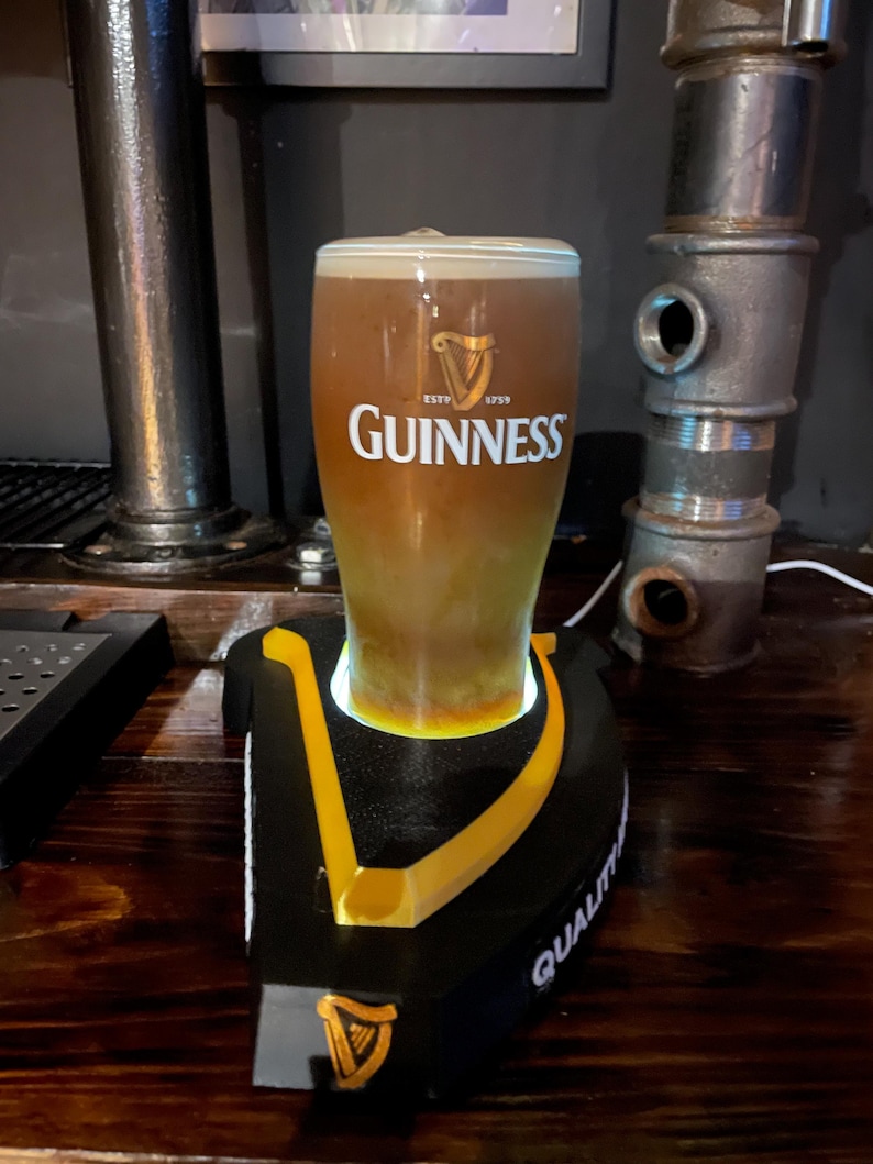 May include: A pint of Guinness stout in a branded glass, resting on a black and yellow branded coaster. The glass is filled with the dark, creamy beer, and the Guinness logo is prominently displayed. The coaster has the word "QUALITY" printed on it.