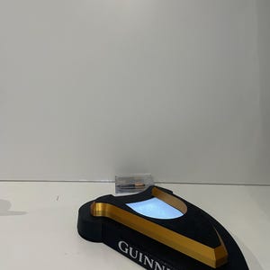 May include: A black and gold Guinness-branded bar accessory with a rectangular base and a curved top. The word "GUINNESS" is printed in white on the front. The top has a light-up feature.