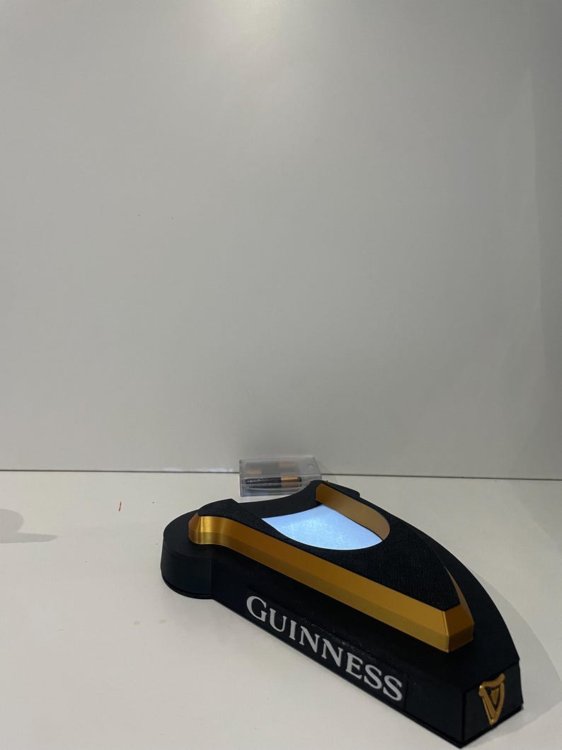 May include: A black and gold Guinness-branded bar accessory with a rectangular base and a curved top. The word "GUINNESS" is printed in white on the front. The top has a light-up feature.