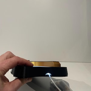 May include: A black and gold display stand with a bright white light. The stand has a rectangular base and a gold-colored top. A clear plastic battery pack is connected to the stand.