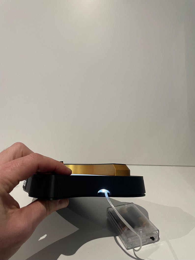May include: A black and gold display stand with a bright white light. The stand has a rectangular base and a gold-colored top. A clear plastic battery pack is connected to the stand.