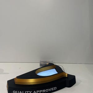 May include: A black and gold display stand with the text "QUALITY APPROVED". The stand has a curved design with a light-up interior. The stand is on a white surface.