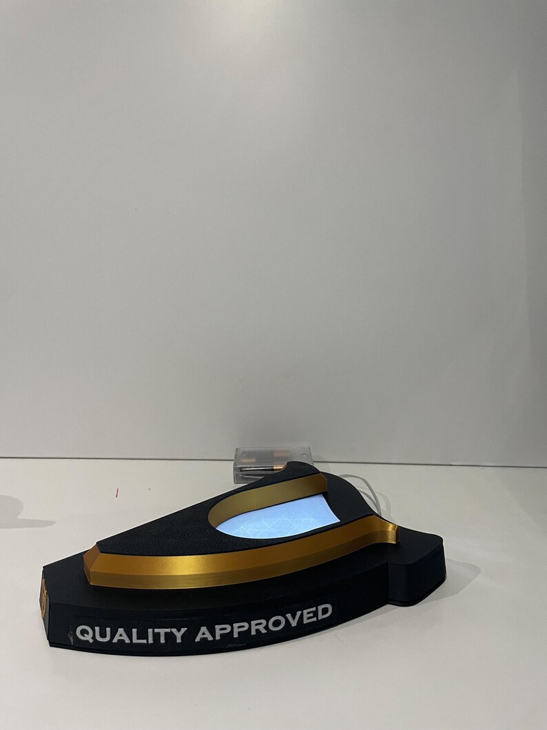 May include: A black and gold display stand with the text "QUALITY APPROVED". The stand has a curved design with a light-up interior. The stand is on a white surface.
