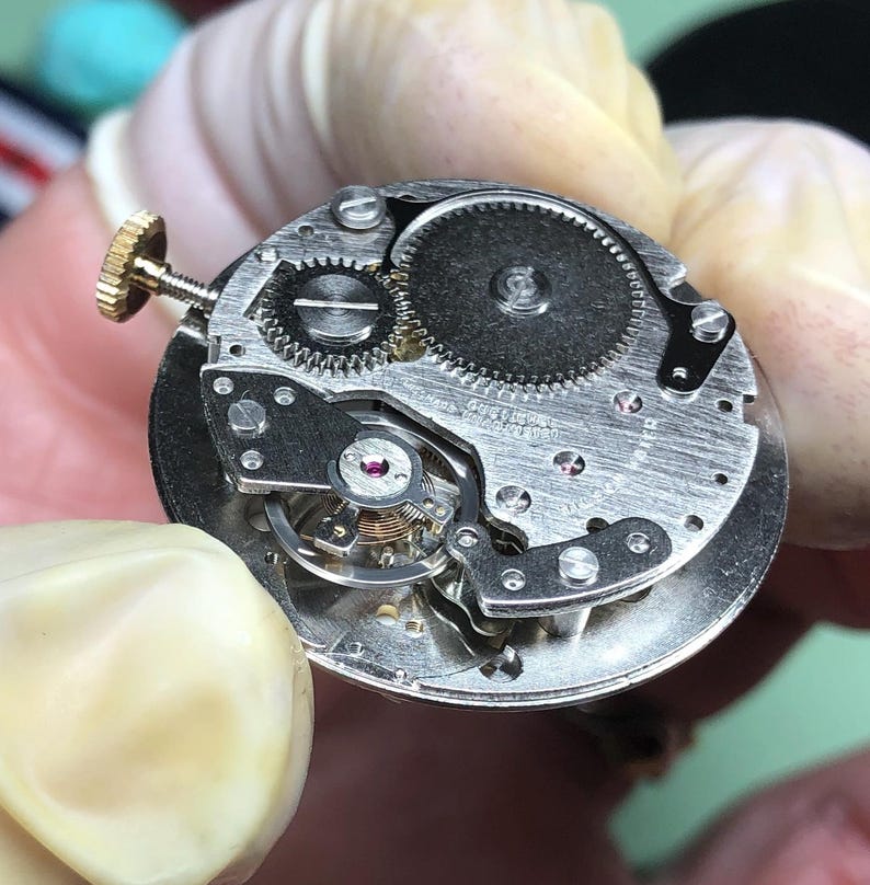 May include: Close-up of a watch movement, showcasing intricate gears, screws, and a balance wheel. The metal components are silver-toned, with a gold-colored winding stem. The design reveals the inner workings of a mechanical timepiece.