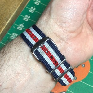 May include: A striped watch band in navy blue, white, and red, with a silver-colored buckle. The band is wrapped around a wrist, showcasing the woven fabric and the metal hardware.