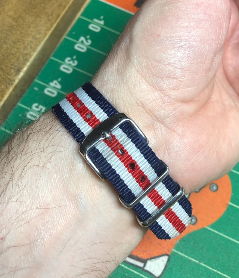 May include: A striped watch band in navy blue, white, and red, with a silver-colored buckle. The band is wrapped around a wrist, showcasing the woven fabric and the metal hardware.