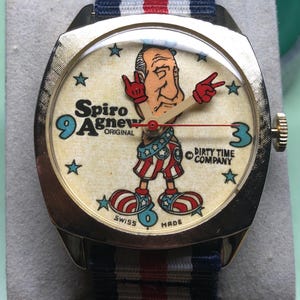 May include: Vintage gold-tone wristwatch with a cream-colored face featuring a cartoon figure and the text "Spiro Agnew Original." The watch has a striped blue, white, and red band. The watch face also has the text "Dirty Time Company" and "Swiss Made."