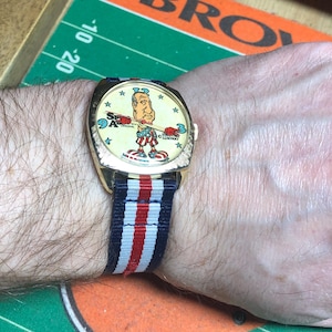 May include: A vintage wristwatch with a gold-toned case and a colorful cartoon dial featuring a patriotic figure. The watch has a striped blue, white, and red band. The watch is on a person's wrist.