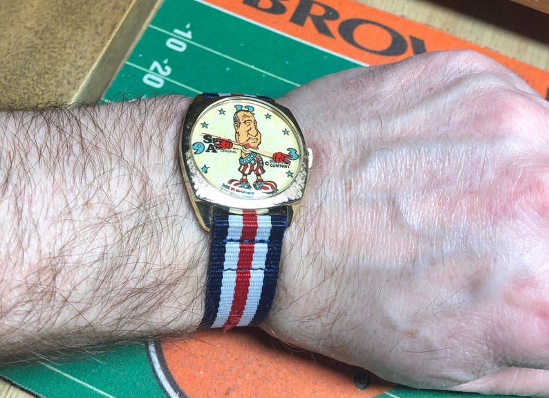 May include: A vintage wristwatch with a gold-toned case and a colorful cartoon dial featuring a patriotic figure. The watch has a striped blue, white, and red band. The watch is on a person's wrist.