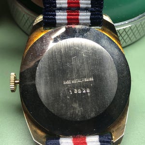 May include: A vintage wristwatch with a gold-toned case and a navy, white, and red striped band. The watch back is silver with the text "BASE METAL SWISS" and the number "18826". The watch is on a green surface.