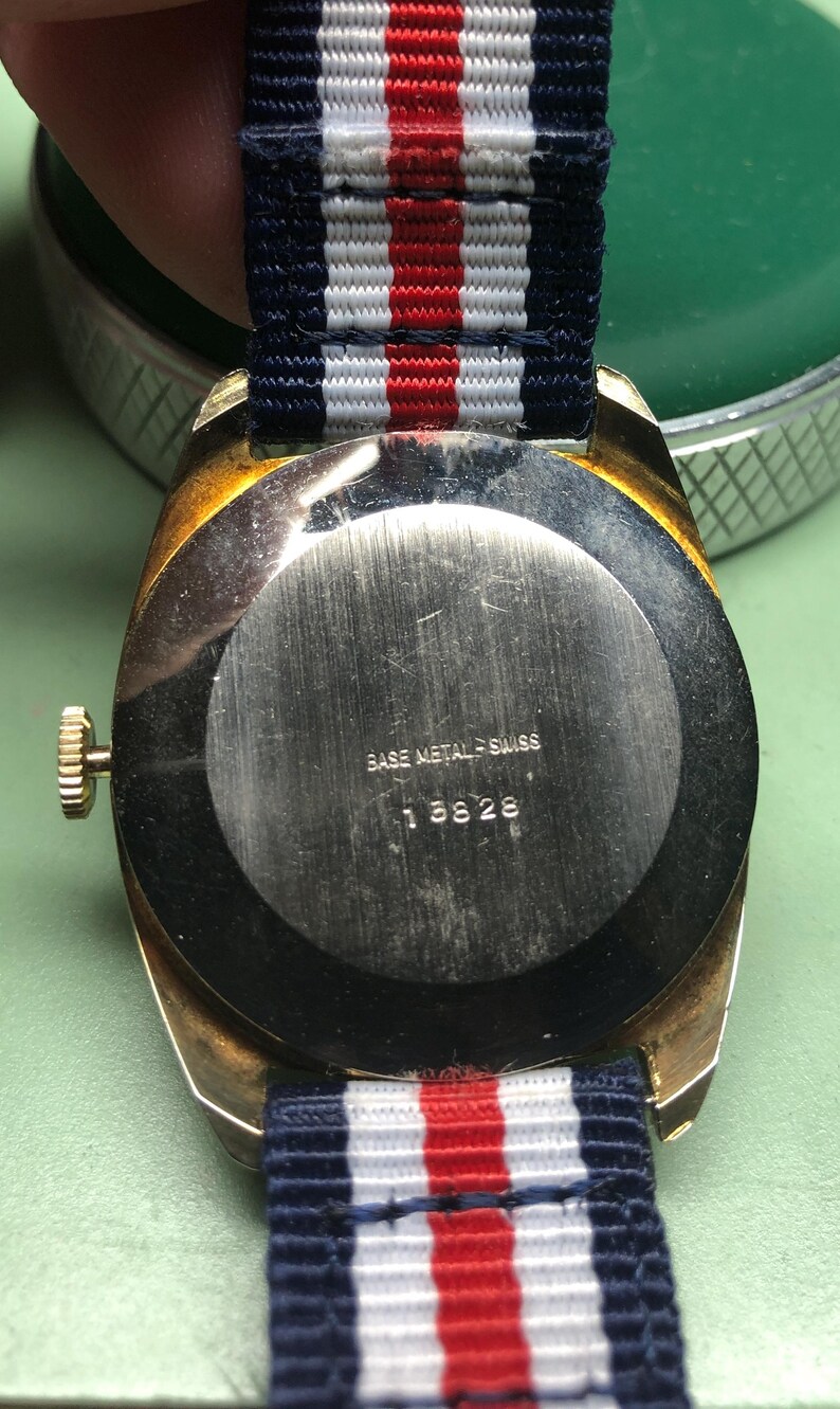 May include: A vintage wristwatch with a gold-toned case and a navy, white, and red striped band. The watch back is silver with the text "BASE METAL SWISS" and the number "18826". The watch is on a green surface.