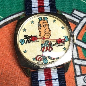 May include: Vintage wristwatch with a cream-colored face featuring a cartoon character and the text "Spin 9A Original". The watch has a gold-tone case and a striped navy, red, and white band.