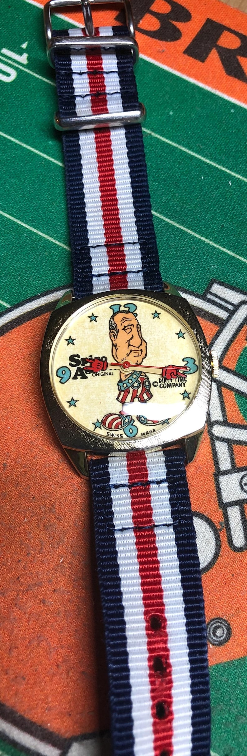 May include: Vintage wristwatch with a cream-colored face featuring a cartoon character and the text "Spin 9A Original". The watch has a gold-tone case and a striped navy, red, and white band.