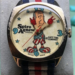 May include: Gold-tone wristwatch with a cream-colored face featuring a cartoon figure and the text "Spiro Agnew ORIGINAL". The watch has a blue, white, and red striped band. The watch face also has the text "DIRTY TIME COMPANY".