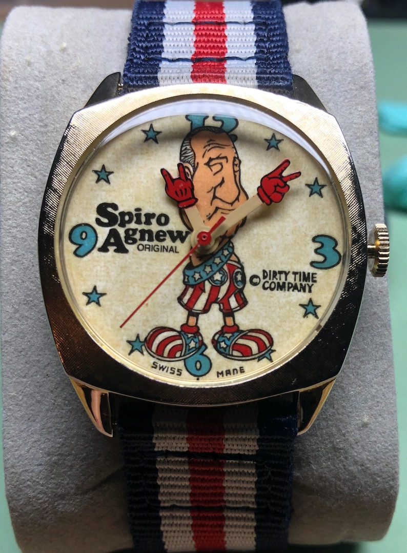 May include: Gold-tone wristwatch with a cream-colored face featuring a cartoon figure and the text "Spiro Agnew ORIGINAL". The watch has a blue, white, and red striped band. The watch face also has the text "DIRTY TIME COMPANY".