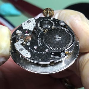 May include: Close-up of a vintage Wilson Watch movement. The silver-toned mechanism features intricate gears, a balance wheel with a red jewel, and the text "WILSON WATCH". The watch movement is held by a gloved hand.