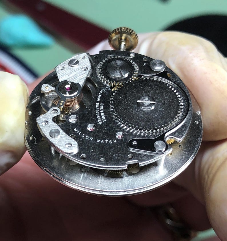 May include: Close-up of a vintage Wilson Watch movement. The silver-toned mechanism features intricate gears, a balance wheel with a red jewel, and the text "WILSON WATCH". The watch movement is held by a gloved hand.