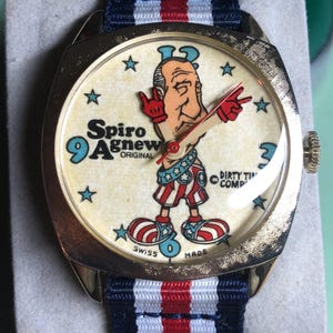 May include: Vintage gold-toned wristwatch with a cream-colored face featuring a cartoon figure and the text "Spiro Agnew Original". The watch has a blue, red, and white striped band and the text "Swiss Made".