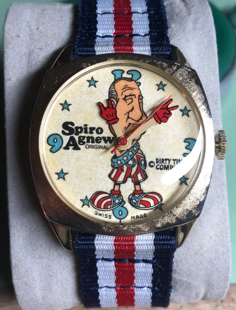 May include: Vintage gold-toned wristwatch with a cream-colored face featuring a cartoon figure and the text "Spiro Agnew Original". The watch has a blue, red, and white striped band and the text "Swiss Made".