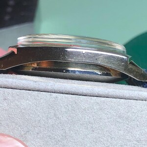 May include: Side view of a vintage wristwatch with a silver-colored case and a clear crystal. The watch has a dark blue fabric strap. The watch is resting on a gray surface.