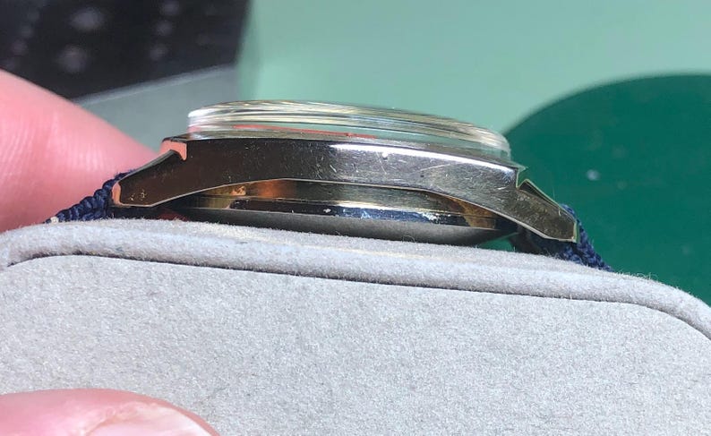 May include: Side view of a vintage wristwatch with a silver-colored case and a clear crystal. The watch has a dark blue fabric strap. The watch is resting on a gray surface.