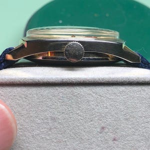 May include: A vintage gold-toned wristwatch with a dark blue fabric band. The watch has a round face with a clear crystal cover. The watch is resting on a gray surface. The watch's crown is visible.