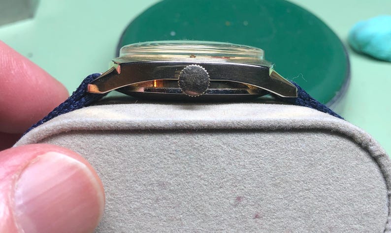 May include: A vintage gold-toned wristwatch with a dark blue fabric band. The watch has a round face with a clear crystal cover. The watch is resting on a gray surface. The watch's crown is visible.