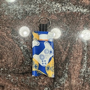 May include: A blue and yellow fabric keychain holder with a video game controller pattern. The holder contains a lip balm tube. The keychain has a silver ring and a black fabric loop.