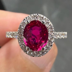 May include: A ring featuring a large, oval-cut, vibrant pinkish-red gemstone set in a silver-toned metal band. The central stone is surrounded by a halo of small, clear, sparkling stones, with additional small stones set along the ring band.