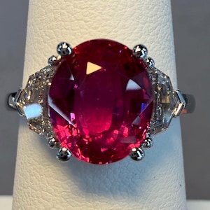 May include: A ring featuring a large, oval-cut, vibrant red gemstone set in a silver-toned metal band. The central red stone is flanked on each side by two smaller, clear, trapezoid-cut stones, all secured by metal prongs on a polished band.