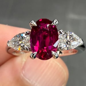 May include: A three-stone ring featuring a central oval-cut deep red ruby held by four prongs. Flanking the center stone are two pear-cut white diamonds. The stones are set on a polished silver-toned metal band with a smooth, reflective finish.